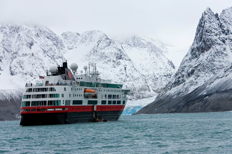 Small expedition cruise ship sailing through remote waters surrounded by dramatic snow-covered mountains.