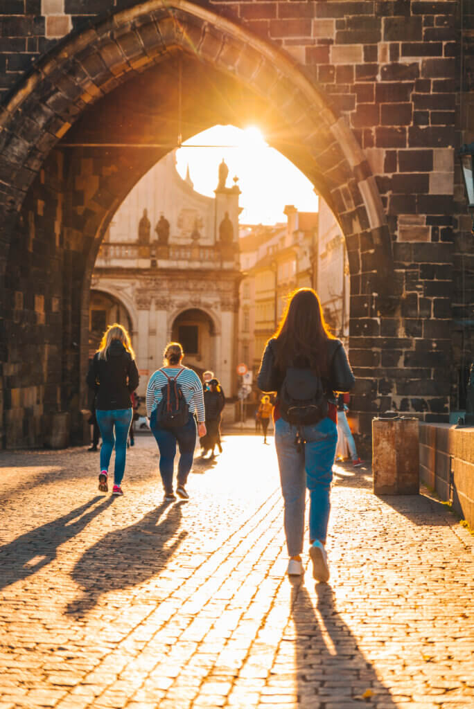 Small group of travelers walking through a historic stone archway at sunset, exploring a cobblestone street and European architecture.