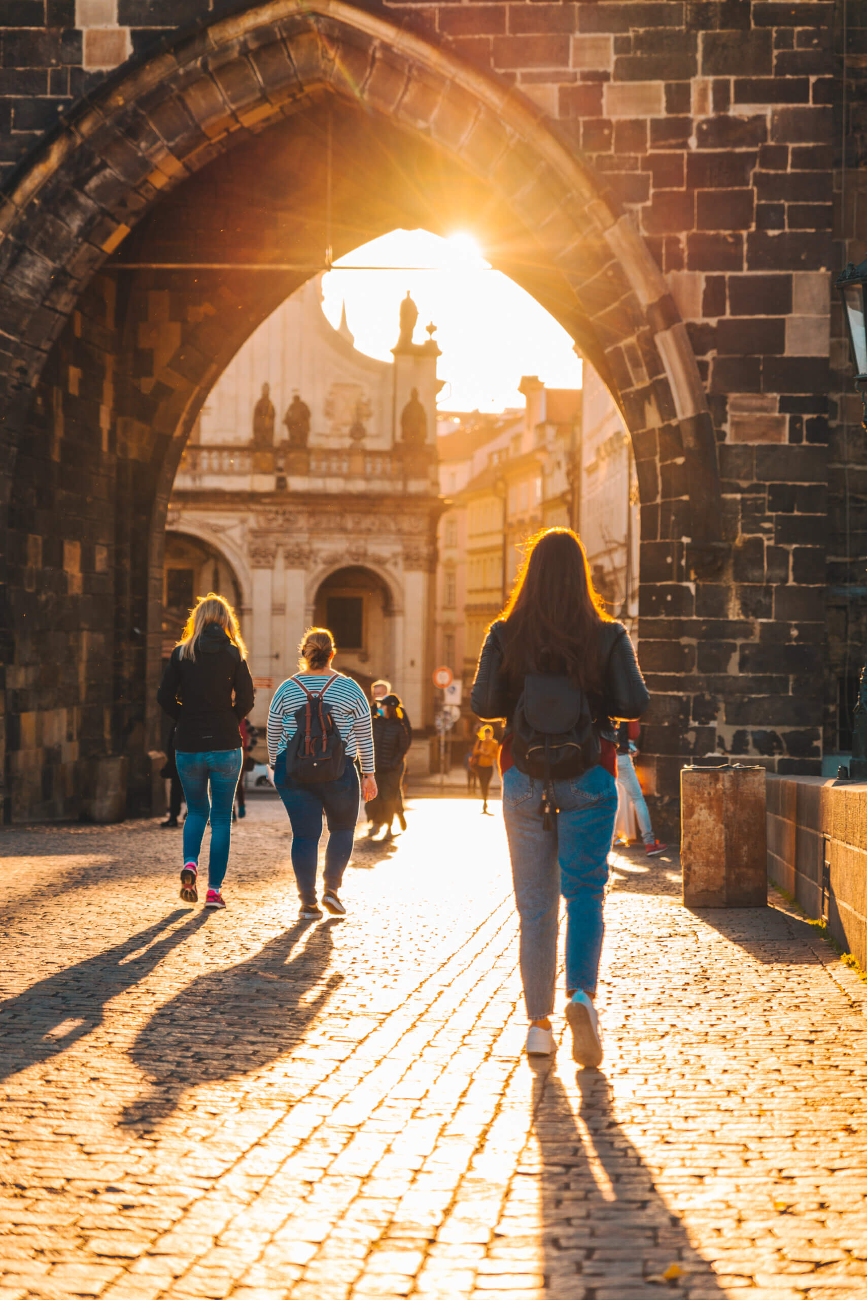 Small group of travelers walking through a historic stone archway at sunset, exploring a cobblestone street and European architecture.
